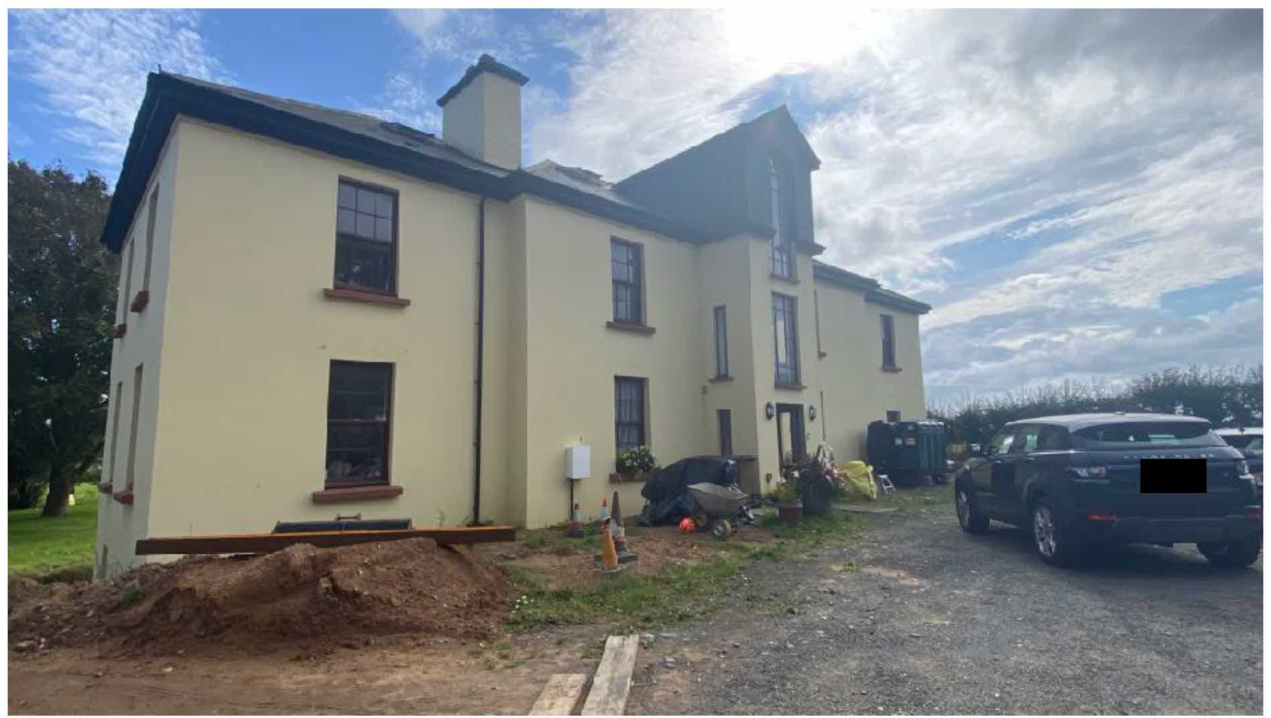 A photograph showing a large, two-story detached house with cream rendering and dark window frames. The foreground features a pile of earth and wooden beams, suggesting landscaping or construction work, with a car par...