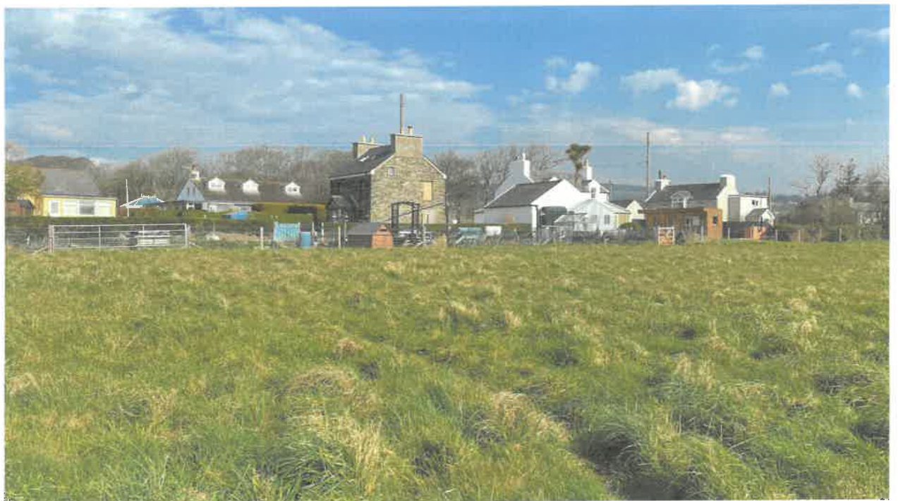 A photograph showing a grassy field in the foreground with a row of residential buildings and a stone structure in the background under a blue sky.