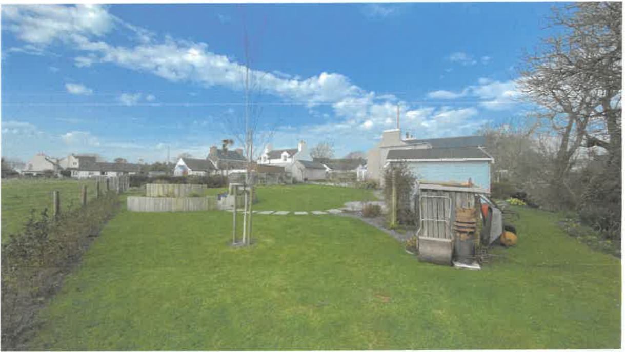 A photograph showing a grassy garden area with a small blue outbuilding and wooden structures, with residential houses visible in the background.