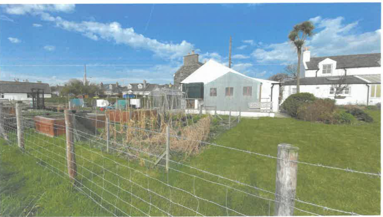 A photograph showing a rural garden area with raised beds and wire fencing in the foreground, with residential buildings and a temporary-looking structure in the background under a blue sky.