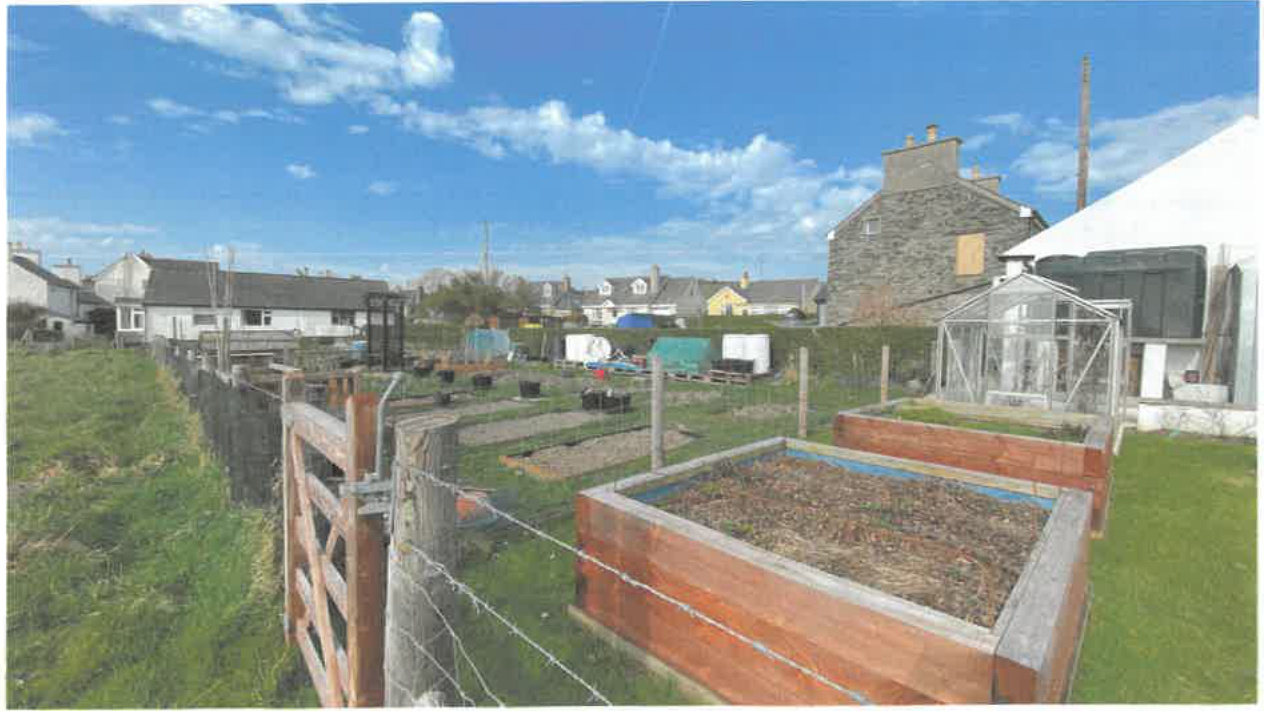 A photograph showing a garden area with raised wooden beds, a greenhouse, and fencing, with stone houses visible in the background.