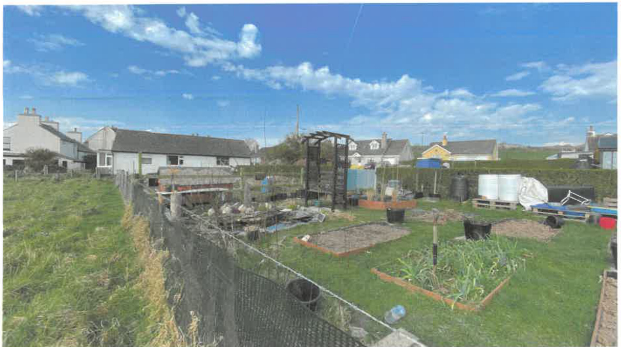 A sunny photograph showing a residential garden with raised vegetable beds and fencing adjacent to a grassy field, with houses visible in the background.