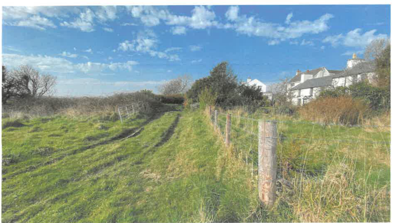A photograph showing a grassy field with a wire fence running along the right side. A white residential building is visible in the background behind vegetation under a blue sky.