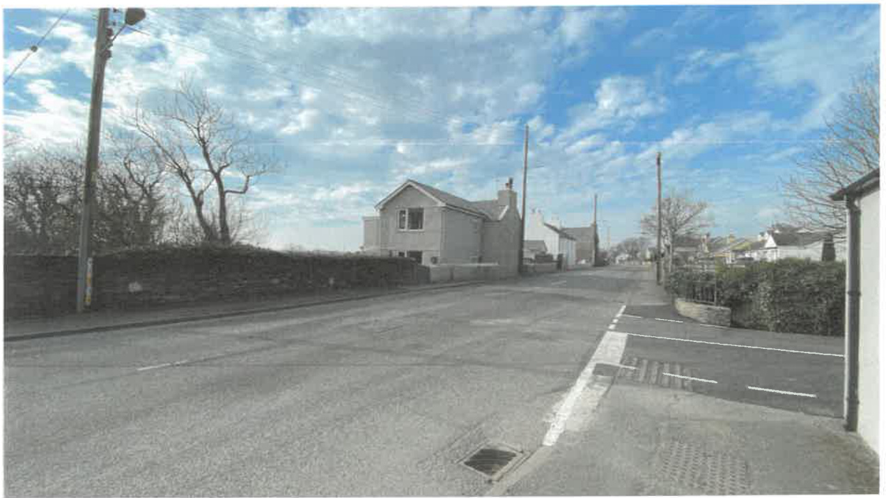 A street-level photograph showing a white detached house on the left side of a road, bordered by a stone wall and trees under a blue sky.