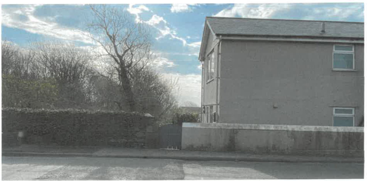 A street-level photograph showing the side elevation of a two-story detached house adjacent to a stone boundary wall and hedge.