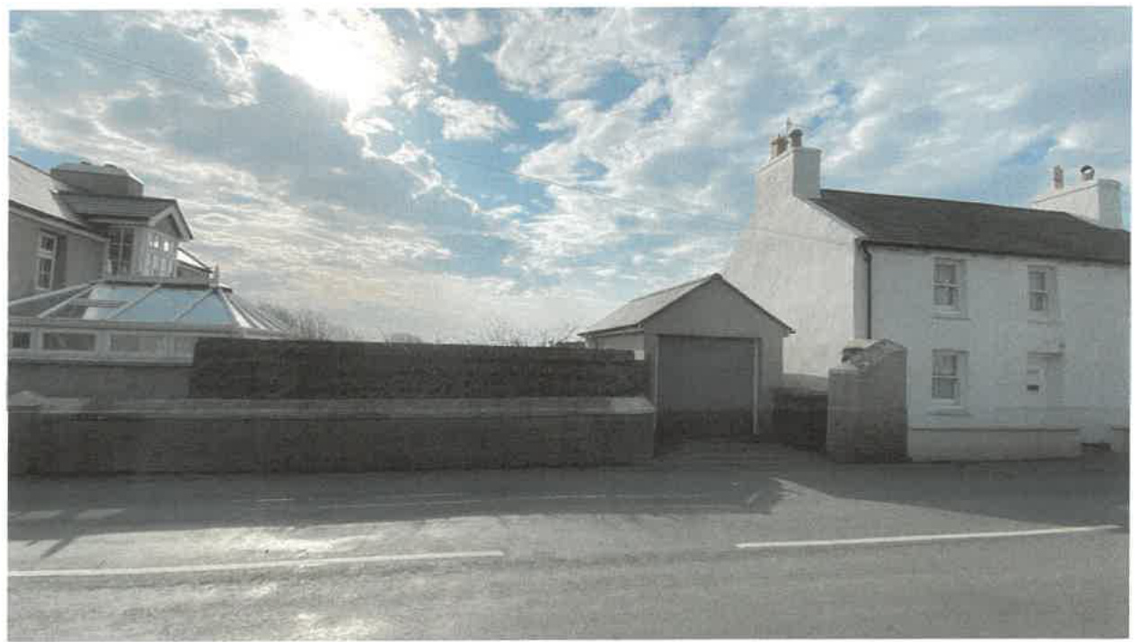A street-level photograph showing a white two-story house on the right, a stone boundary wall, and a small garage or outbuilding in the center.