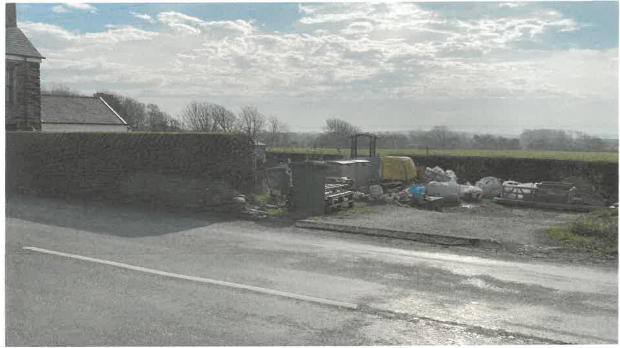 A photograph showing a rural roadside scene with a stone wall boundary, a gravel area containing construction materials and a small excavator, and fields in the background.