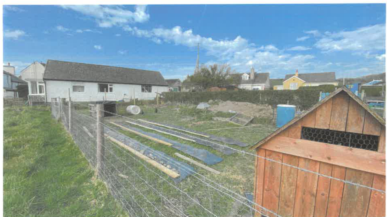 A photograph showing a rural field with a white single-story building and a wooden shed in the foreground, likely depicting the site conditions for a curtilage extension.