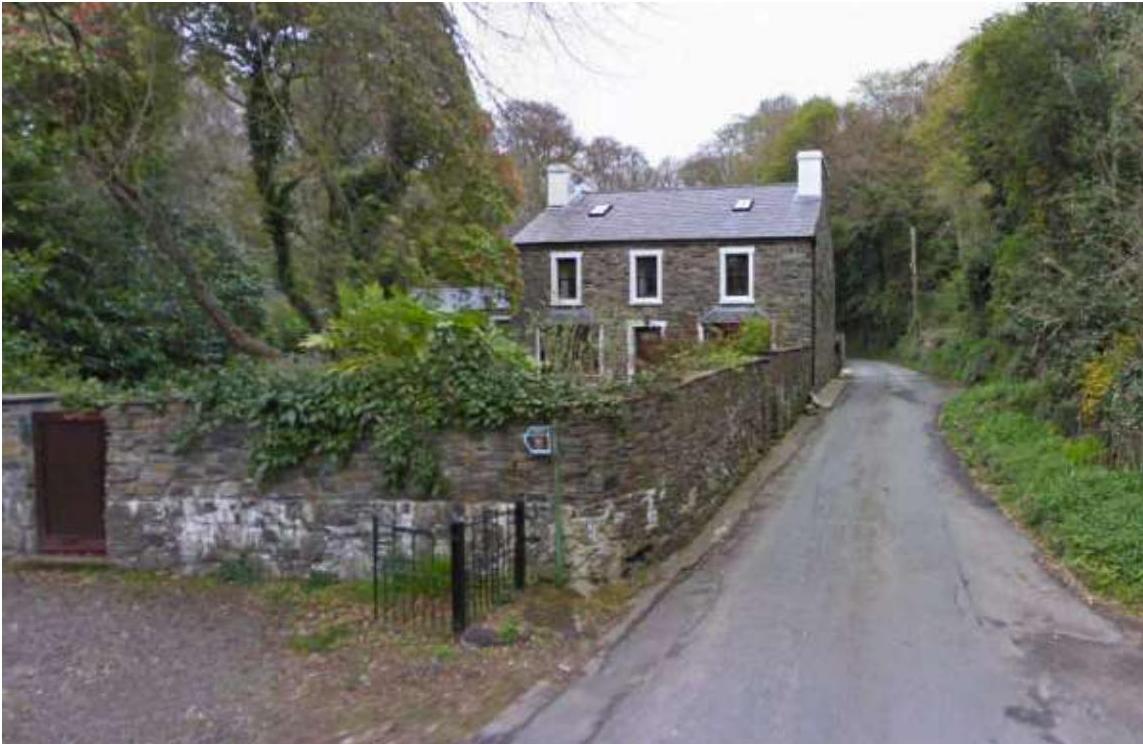 A street-level photograph of a two-story stone house situated on a slope, featuring a stone boundary wall and driveway next to a narrow rural road.