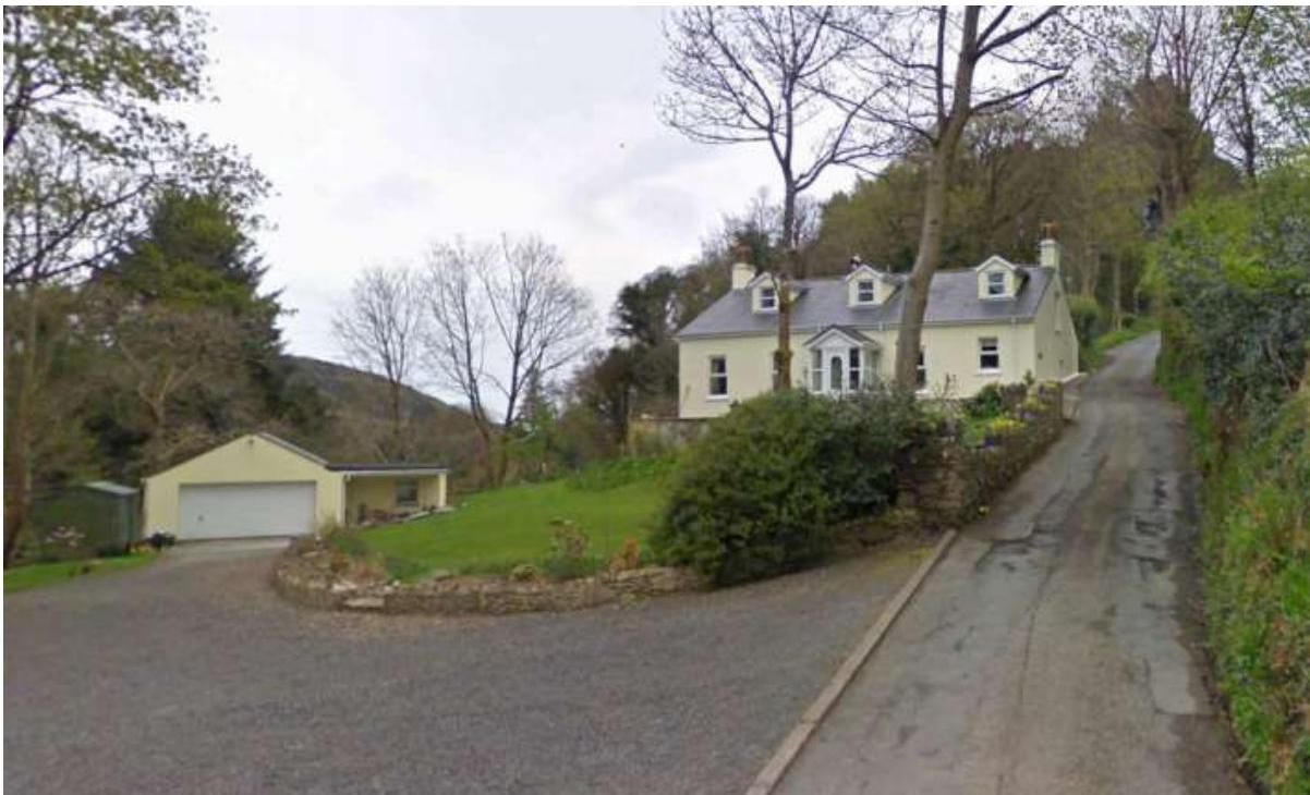 A photograph showing a detached cream house and a separate garage building situated on a sloping rural site with driveway access.