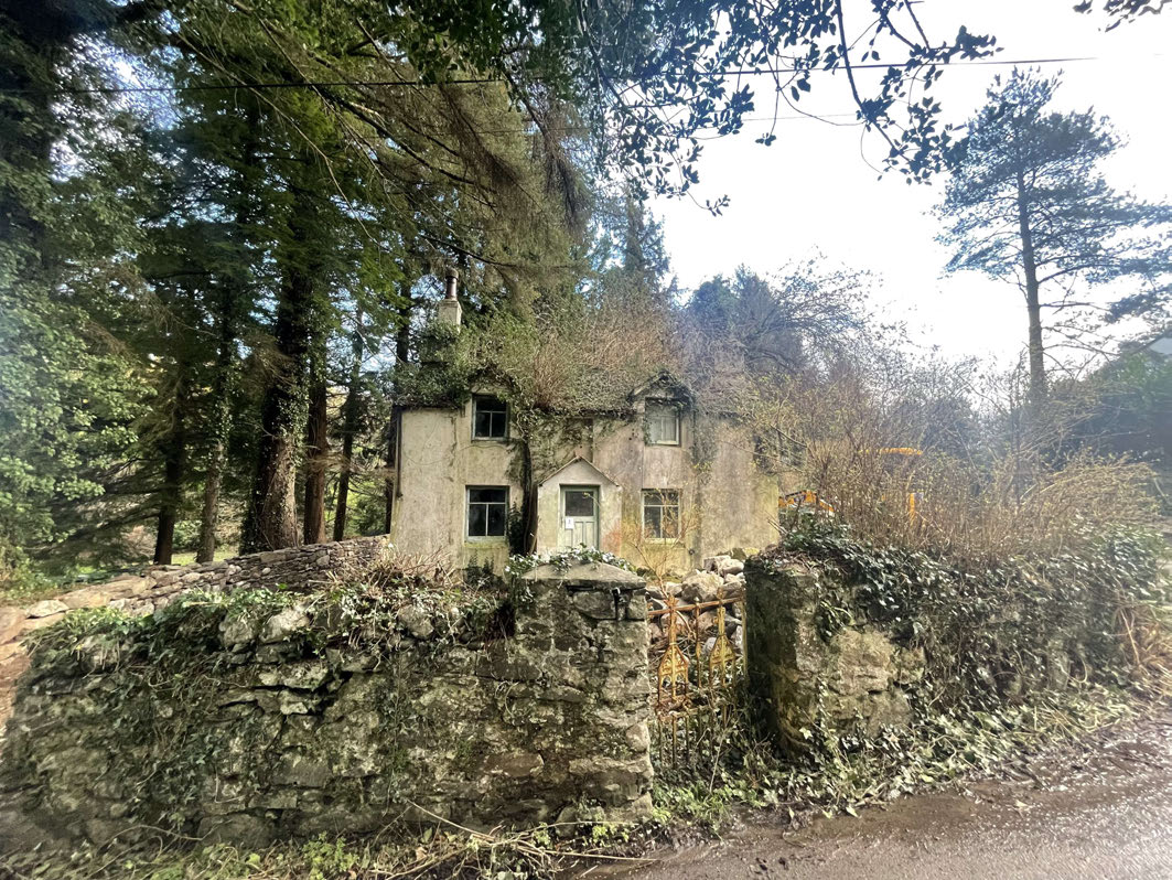 A photograph of a dilapidated, two-story stone cottage surrounded by dense trees and overgrown vegetation, with a stone wall in the foreground.