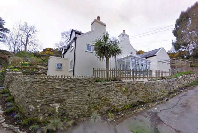 A photograph of a white detached house featuring a conservatory extension, situated behind a prominent stone boundary wall and wooden fence.