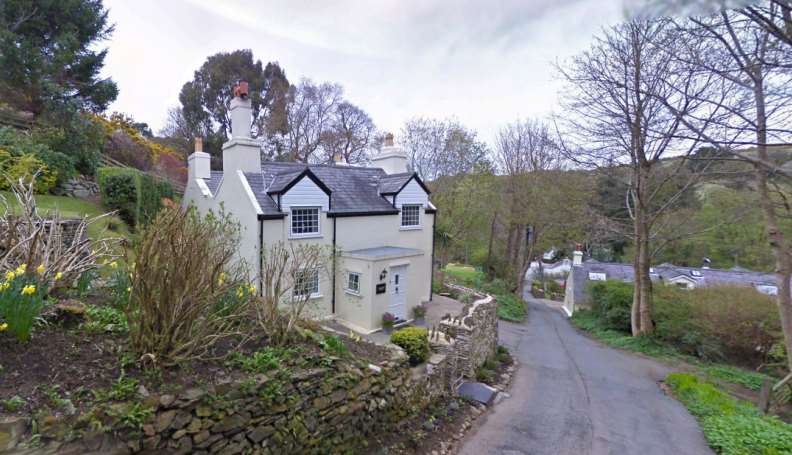 A white detached cottage with a slate roof is situated on a hillside next to a narrow road, surrounded by stone walls and garden vegetation.