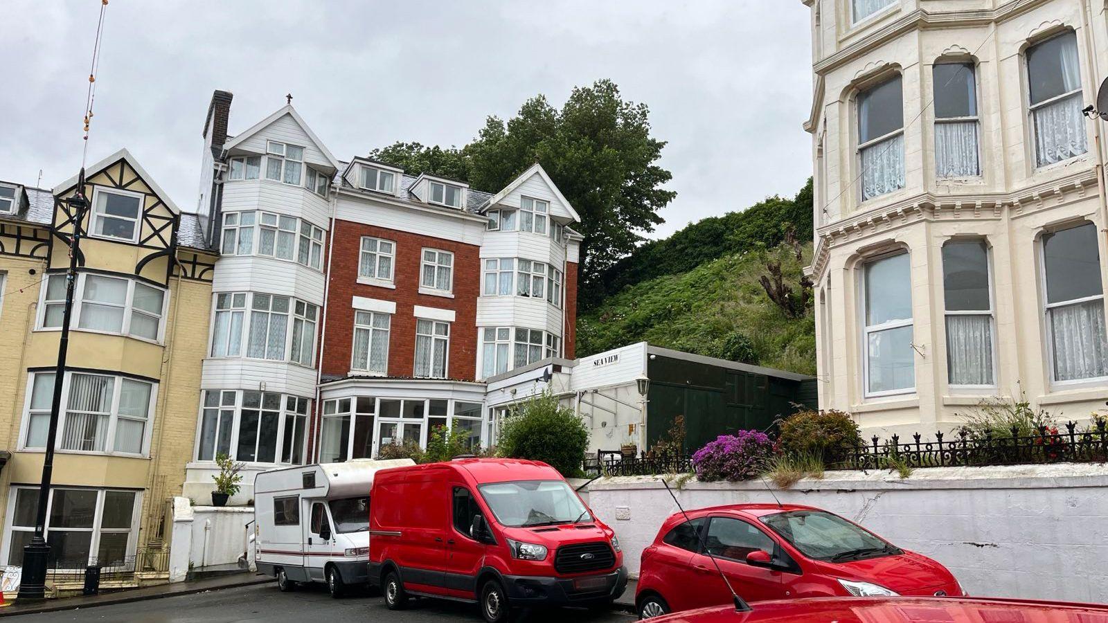 Street-level photograph showing a row of residential buildings in Douglas, including a red brick house and cream-colored terraced properties, with parked vehicles in the foreground.