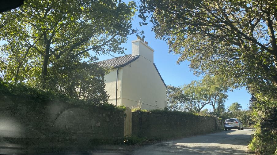 A photograph showing a white detached house viewed through trees, with a stone wall in the foreground and a car parked on the road to the right.