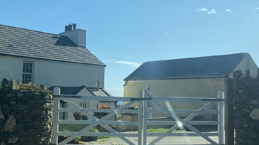 A photograph showing a white detached house with a slate roof on the left and a detached garage or outbuilding on the right, viewed through a white gate and stone wall.