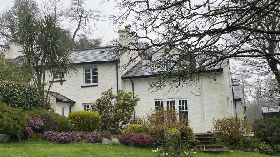 A photograph showing a white, two-story detached house with a slate roof and a garden featuring shrubs and trees.