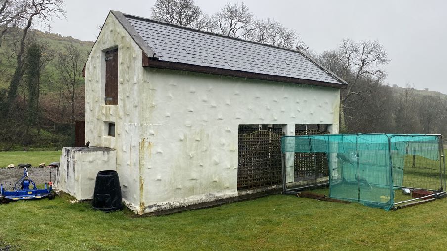 A photograph of a white agricultural building with a slate roof and a green mesh enclosure attached to the side in a rural setting.