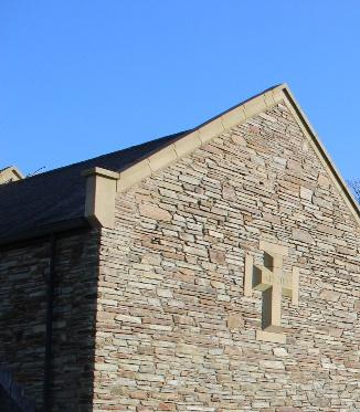 A close-up photograph of the gable end of a stone building featuring a small cross-shaped window or vent against a clear blue sky.