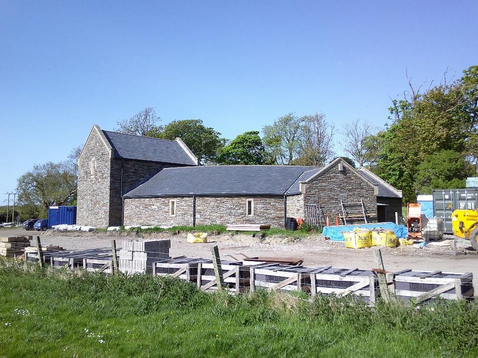 A photograph showing a stone building complex undergoing renovation or construction, with building materials like blocks and bags visible in the foreground.