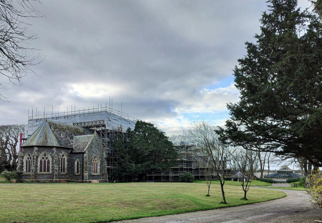 A photograph showing a large stone building covered in scaffolding and white sheeting, likely undergoing renovation. In the foreground, a smaller stone structure with Gothic-style windows sits on a grassy lawn near a...