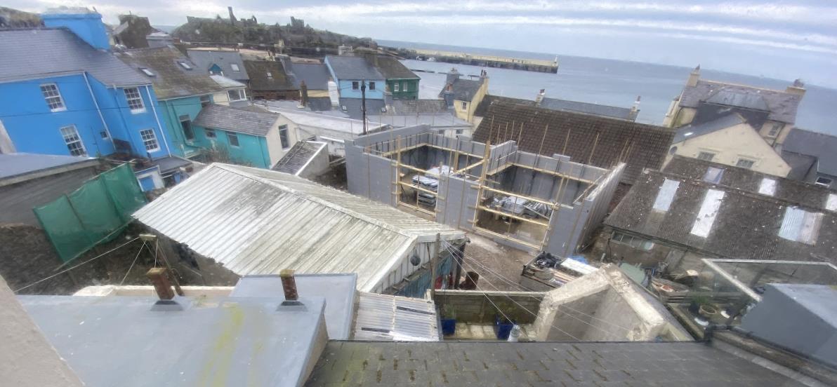 A high-angle photograph showing a construction site with blockwork walls and timber framing, situated in a coastal town with the sea and a pier visible in the background.