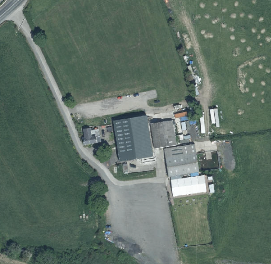 Aerial satellite view of a rural property featuring large industrial or agricultural buildings and gravel parking areas.