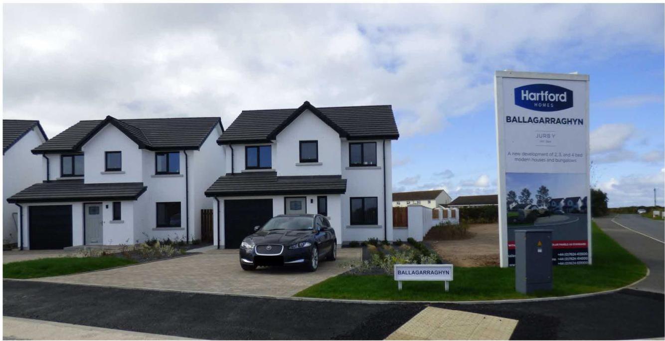 A photograph showing a row of modern white detached houses with dark roofs and a large marketing sign for the Ballagarraghyn development by Hartford Homes.