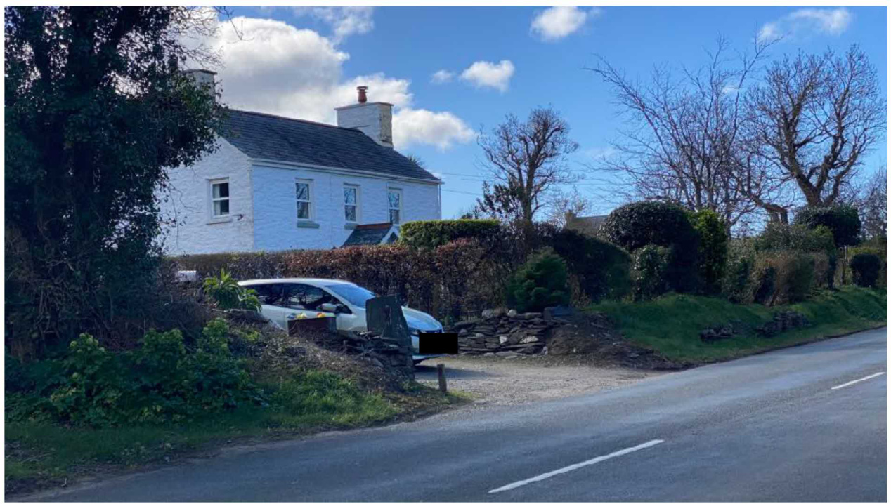 A photograph showing a white two-story detached house with a slate roof situated beside a road, featuring a driveway with a parked car and a stone wall boundary.