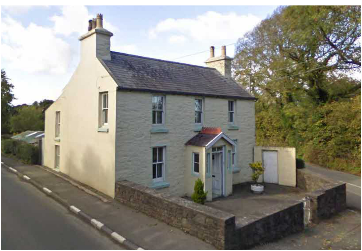 A photograph of a two-story detached stone cottage with a slate roof, sash windows, and a stone boundary wall along a road.