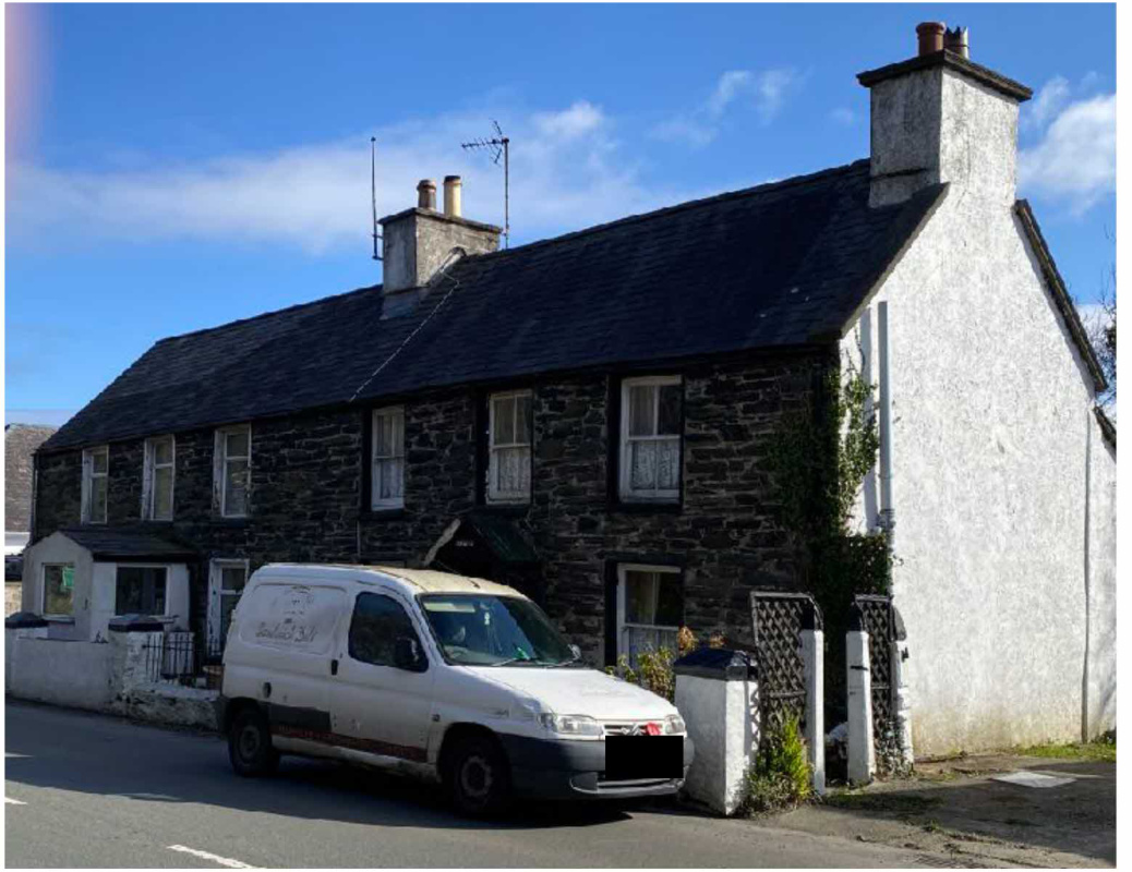 A photograph of a two-story stone and rendered cottage with a slate roof, featuring a white van parked in front on a sunny day.