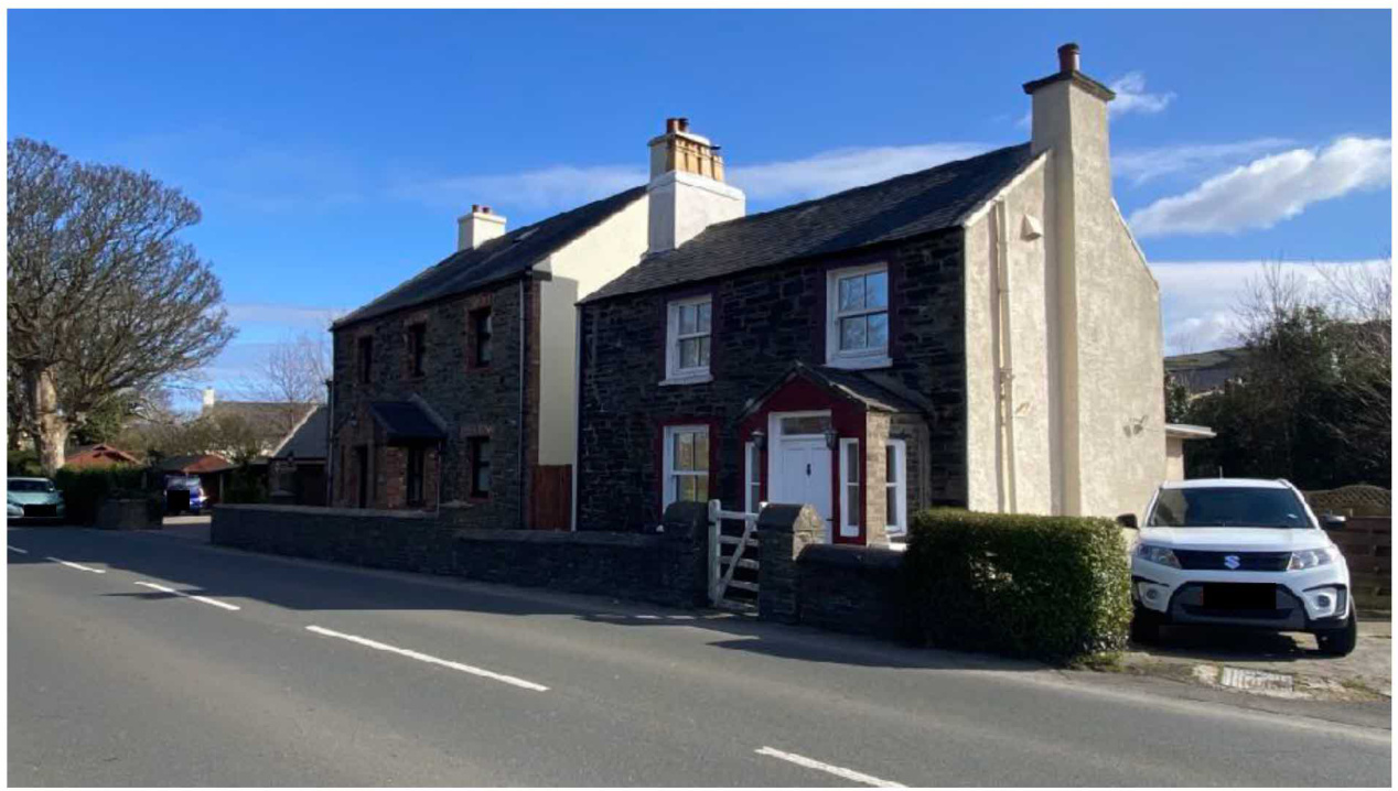 A street-level photograph showing a traditional stone house with white rendered sections and chimneys, situated along a road with a parked car.