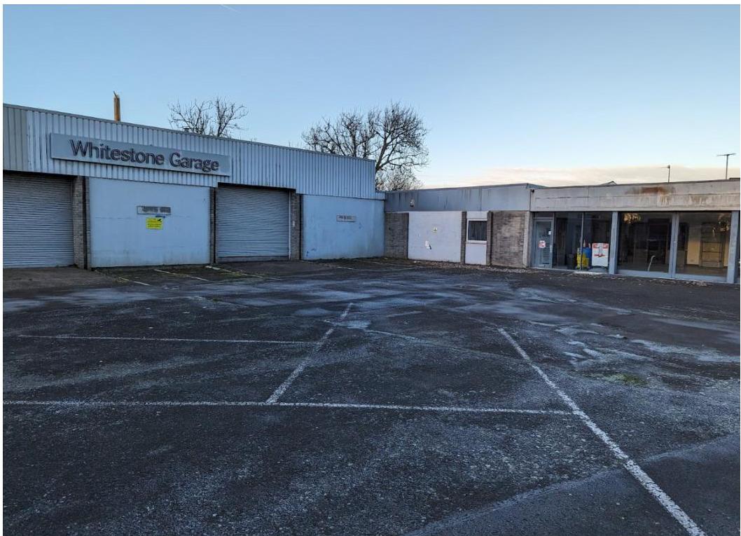 A photograph showing the exterior of a commercial property named 'Whitestone Garage' featuring roller shutter doors and a large paved parking area in the foreground.