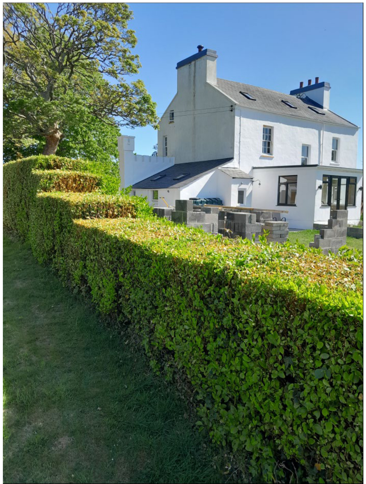 A photograph showing a white detached house with a rear extension and stacks of building blocks on the lawn, viewed from behind a tall hedge.