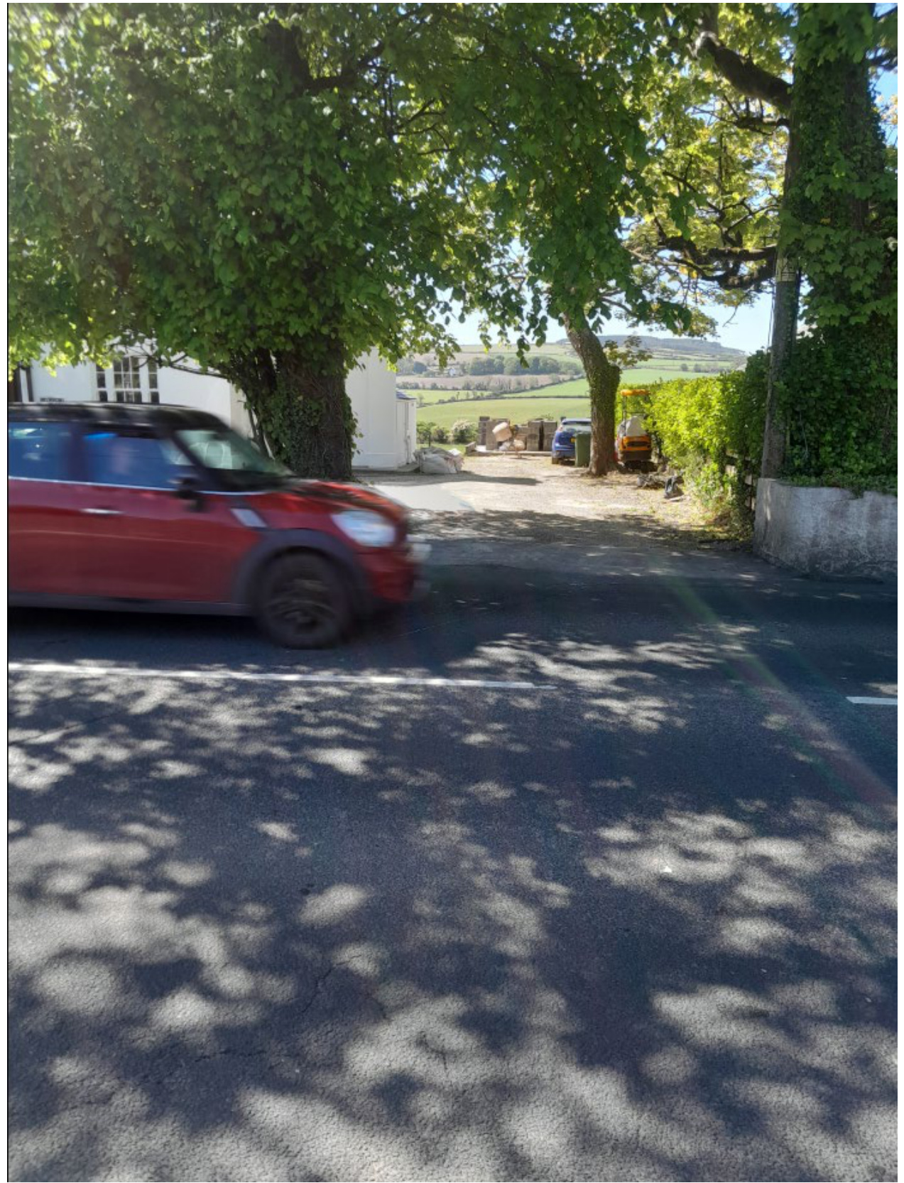 A street-level photograph showing a red car driving past a white building partially obscured by large trees, with rural fields visible in the background.