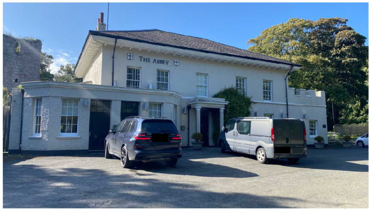 A photograph showing the front exterior of a large white building labeled 'THE ABBEY' with vehicles parked in the driveway.