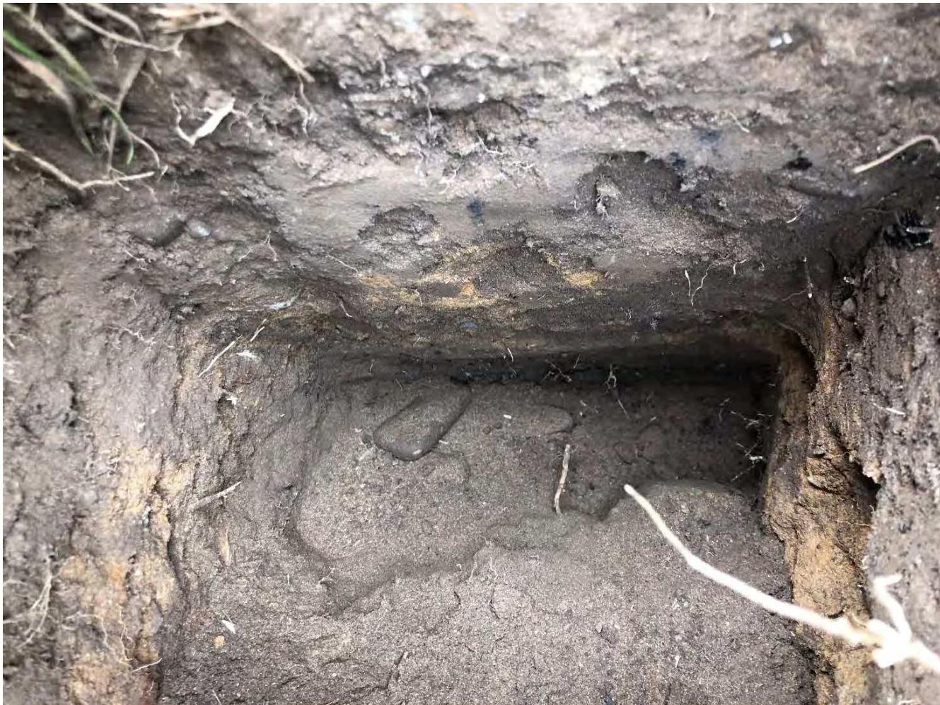 A close-up photograph of an excavated test pit or trial trench showing soil strata and a layer of concrete or hard standing.