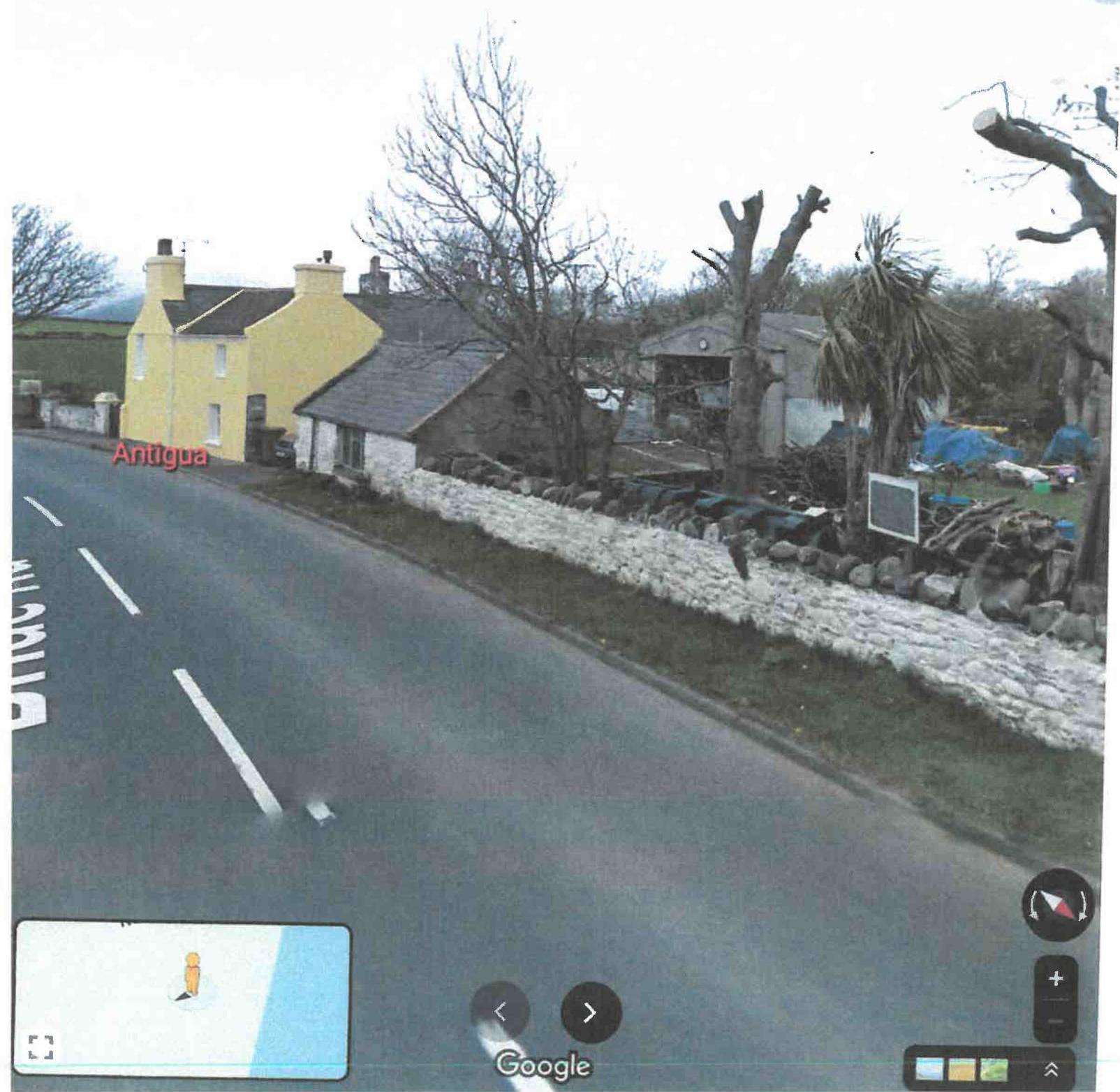 A screenshot from Google Street View showing a rural road with a yellow house, stone buildings, and a garage structure, featuring map interface controls.