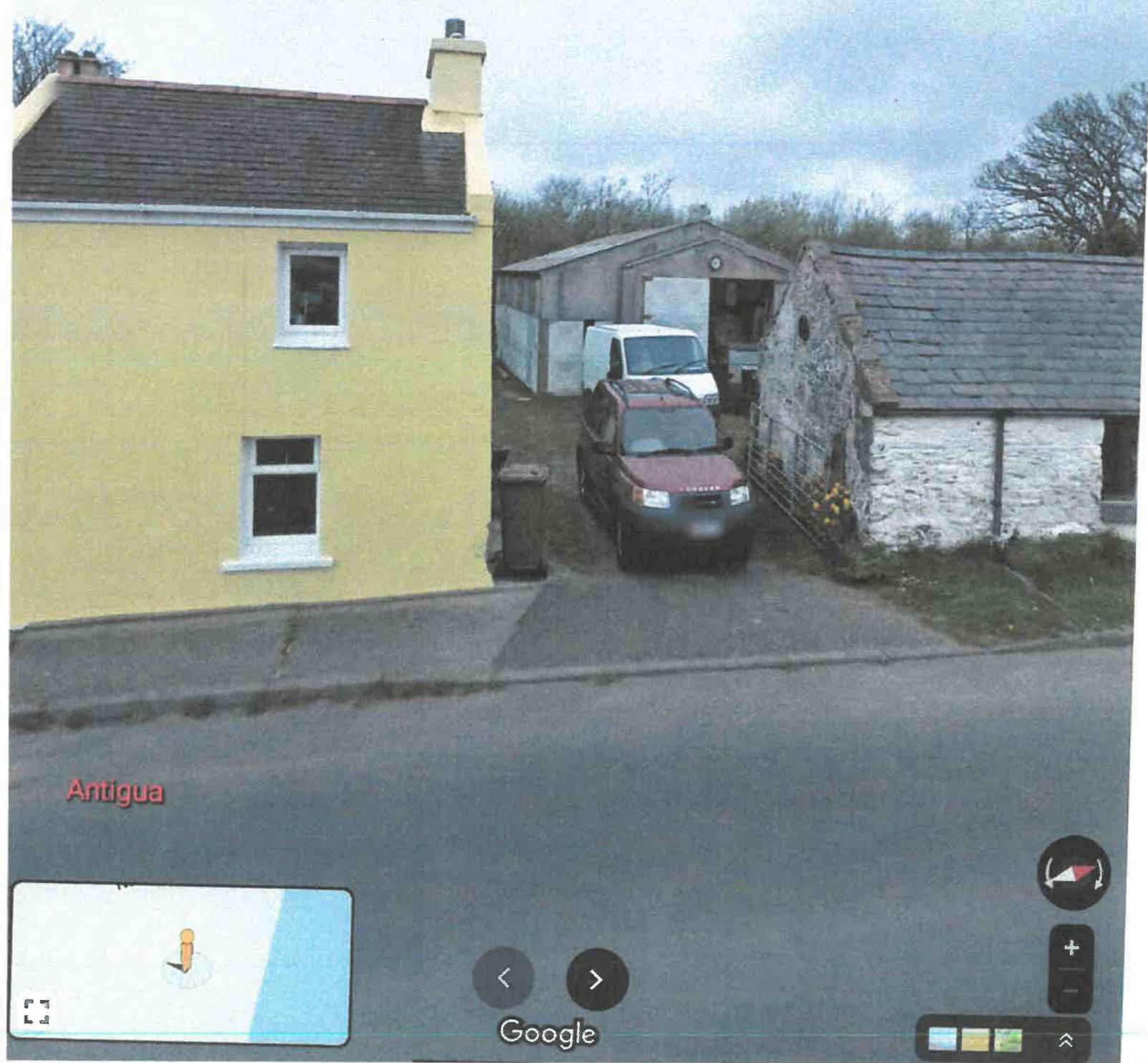 A Google Street View screenshot showing a yellow two-story house, a driveway with parked vehicles, and an adjacent stone outbuilding.