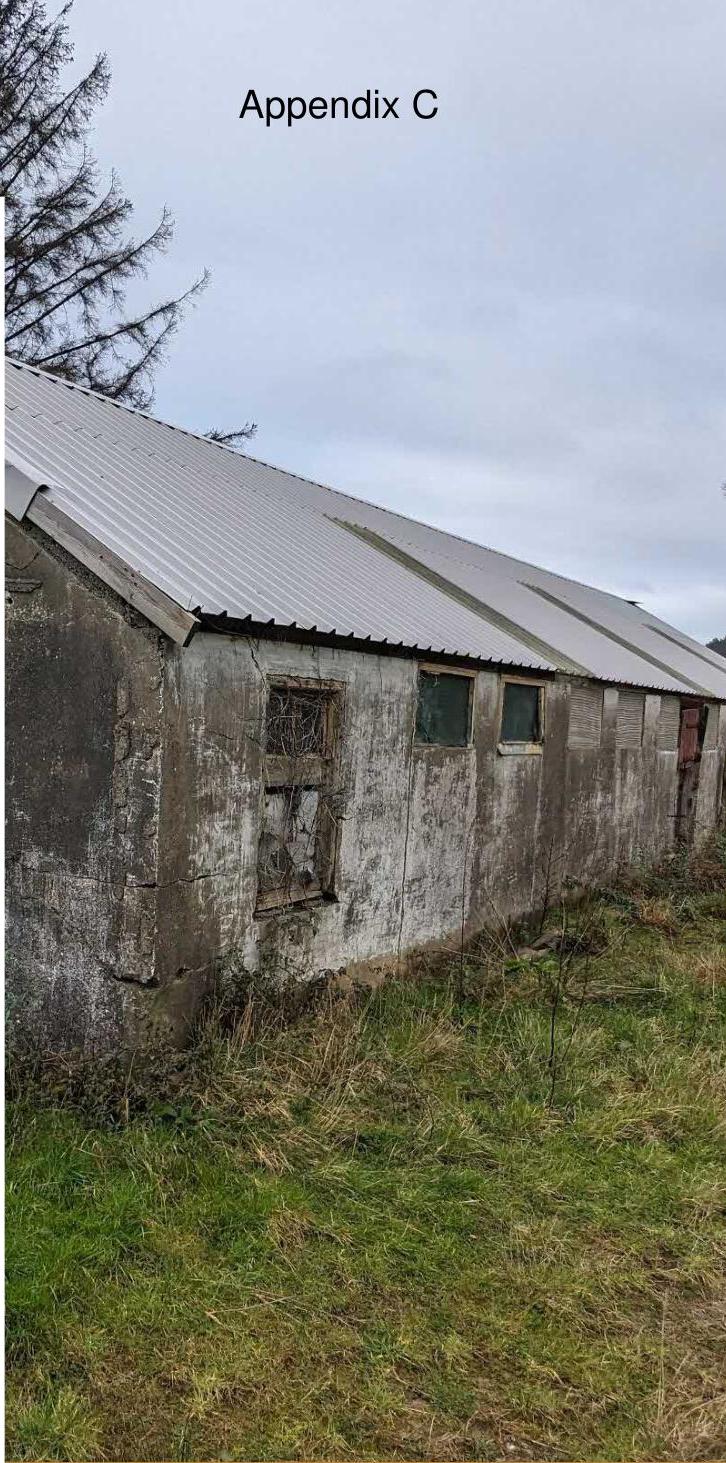 A photograph showing the side elevation of a long, weathered agricultural barn with a corrugated metal roof and concrete walls.