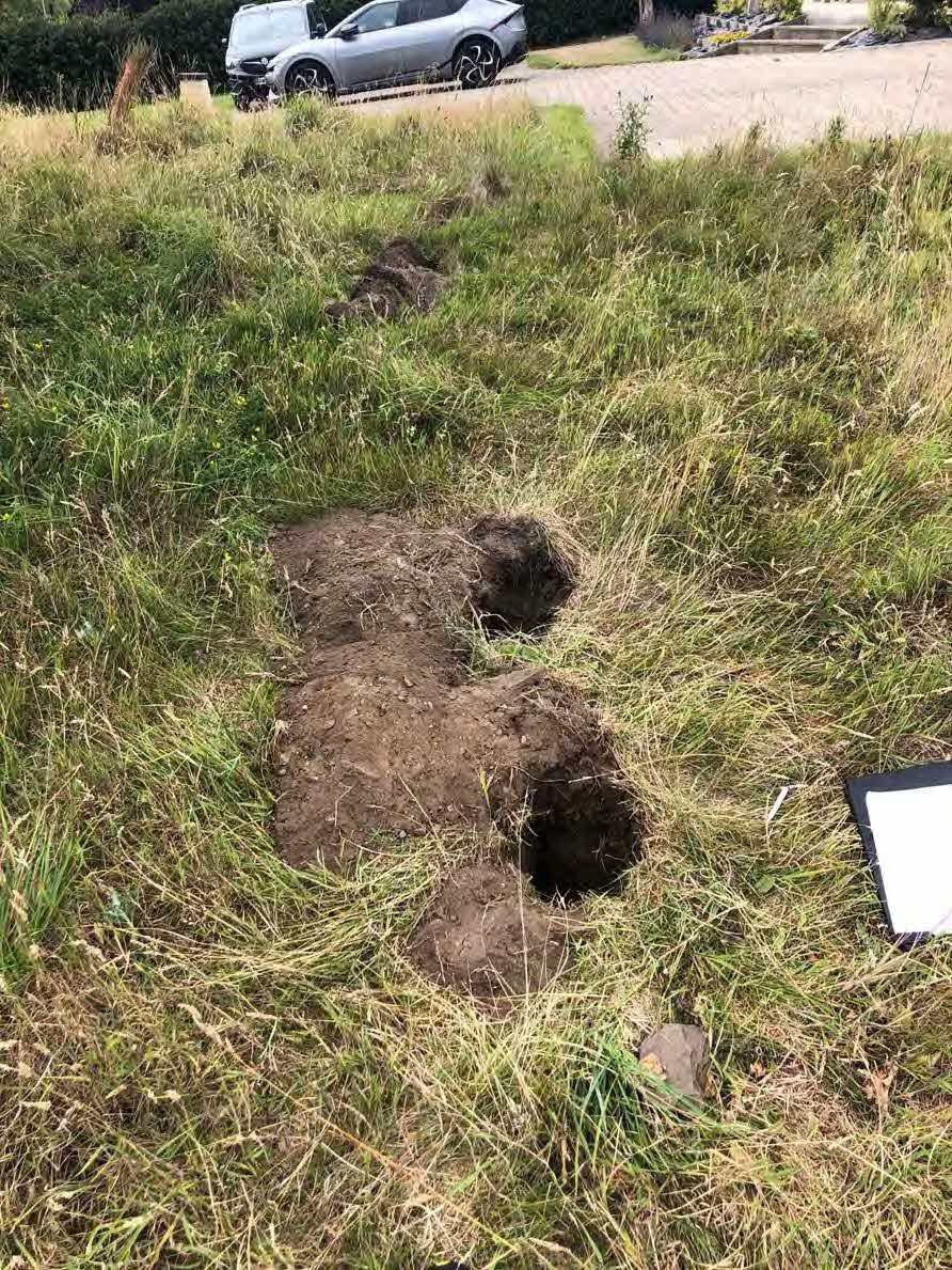 A photograph showing several excavated test pits or boreholes dug into a grassy area, likely for a ground investigation, with a car parked on a driveway in the background.