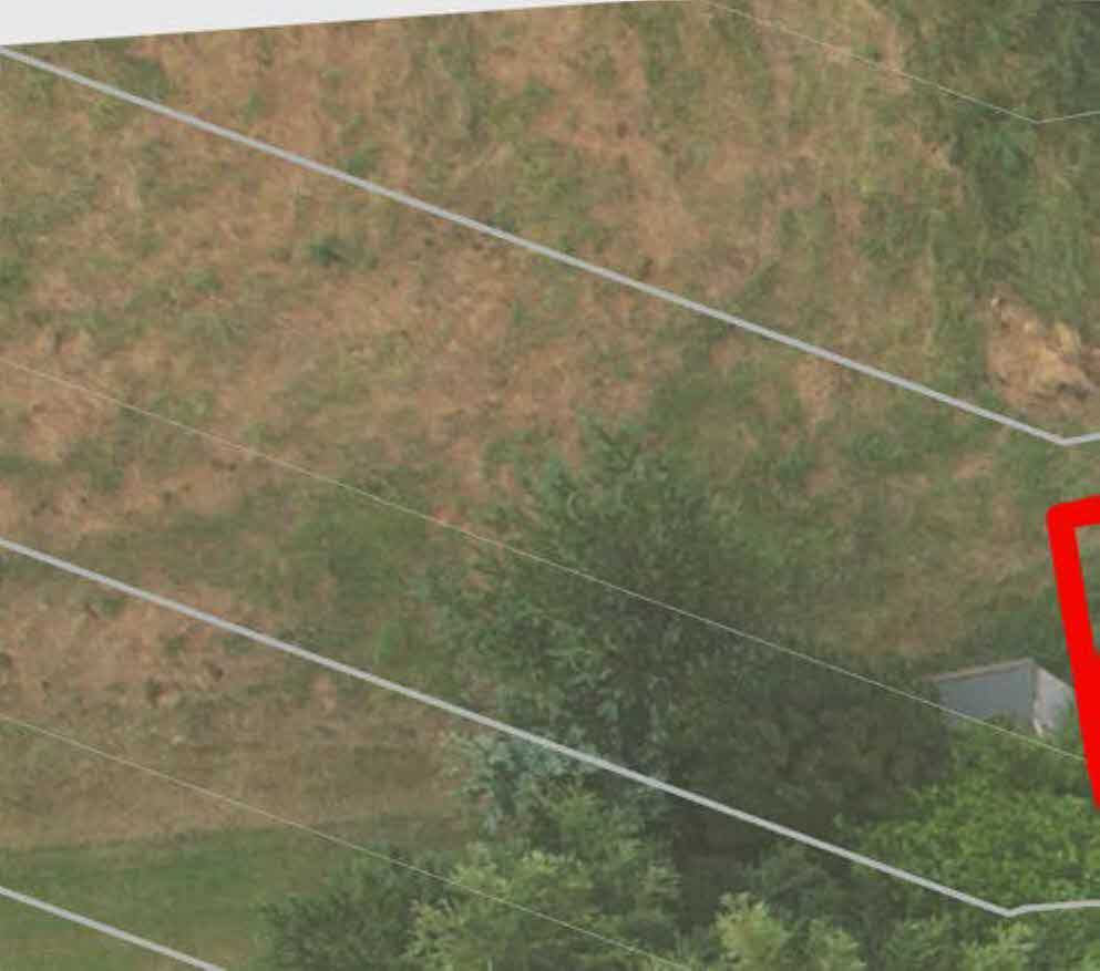 An aerial view of a grassy, sloping plot of land with white boundary lines overlaid, showing a small outbuilding in the corner.