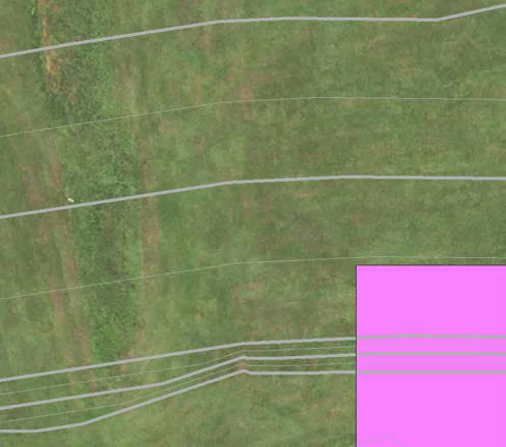An aerial view of a grassy field with faint fence lines and a bright pink highlighted area in the bottom right corner indicating a site boundary.