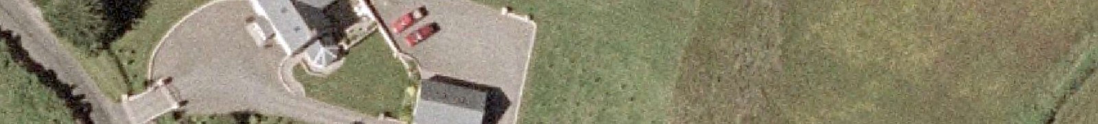 An aerial view of a rural property showing a detached house, a driveway with two red cars, and a separate garage structure surrounded by grassy fields.