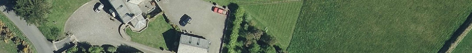 An aerial view showing a rural property with a detached house, a large gravel driveway with parked cars, and adjacent green agricultural fields.