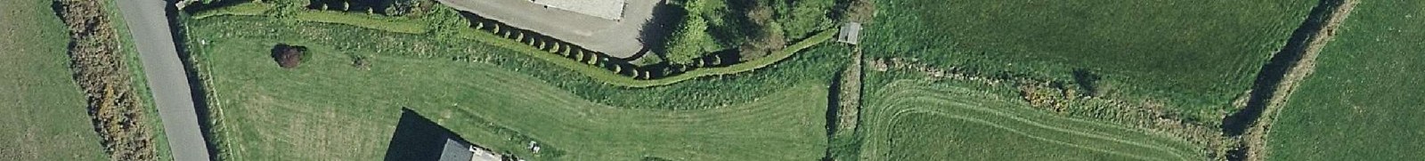 An aerial view of a rural property showing a road, green fields, hedges, and the roof of a building.