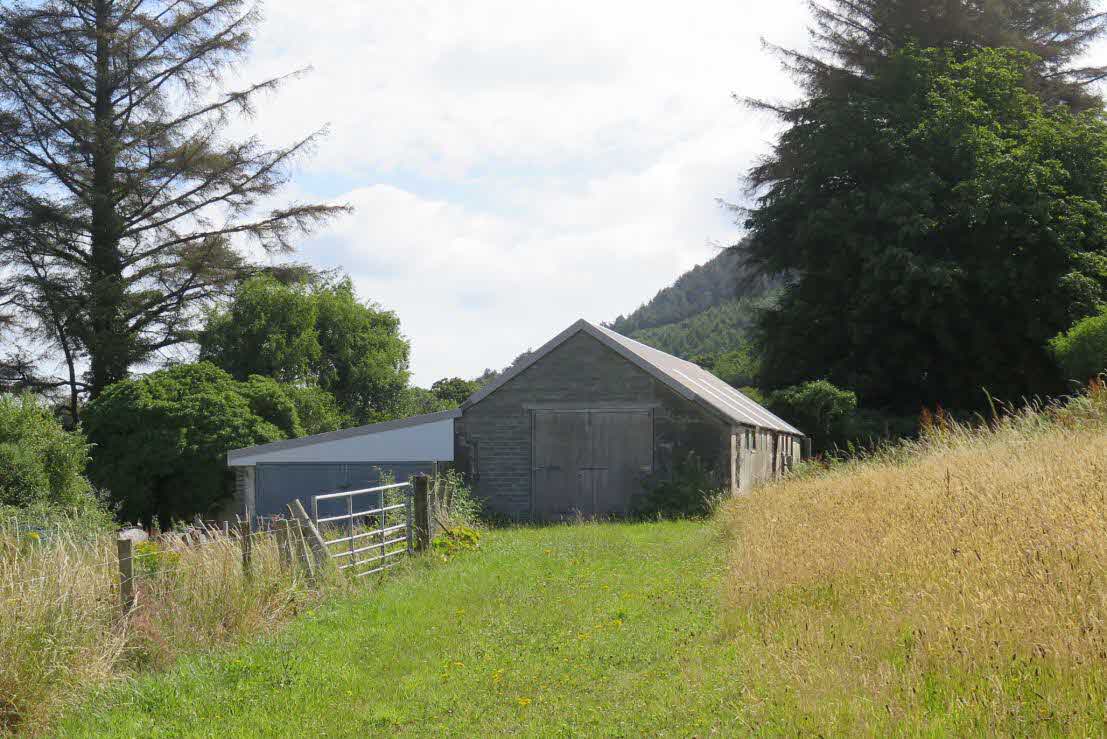 A photograph showing a large, grey agricultural barn situated in a grassy rural field with trees and fencing nearby.