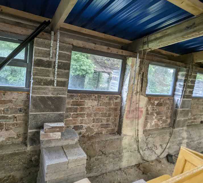 Interior photograph of a building under renovation featuring exposed brick walls, newly installed windows, and wooden roof beams with blue corrugated sheeting.