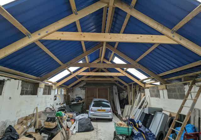 Interior view of a large barn or outbuilding featuring exposed wooden roof trusses and blue corrugated roofing, with a car parked inside.