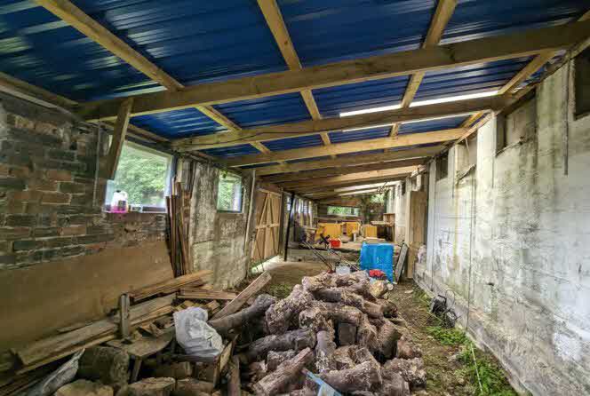 Interior photograph of a long, narrow barn structure with a blue corrugated roof and a large pile of logs on the floor.
